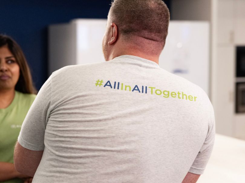 An Alliant Credit Union speaks to a colleague in an office while wearing a company-branded T-shirt that reads #AllInAllTogether